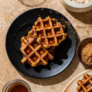 Two small round waffles on a black plate with more waffles and toppings around.