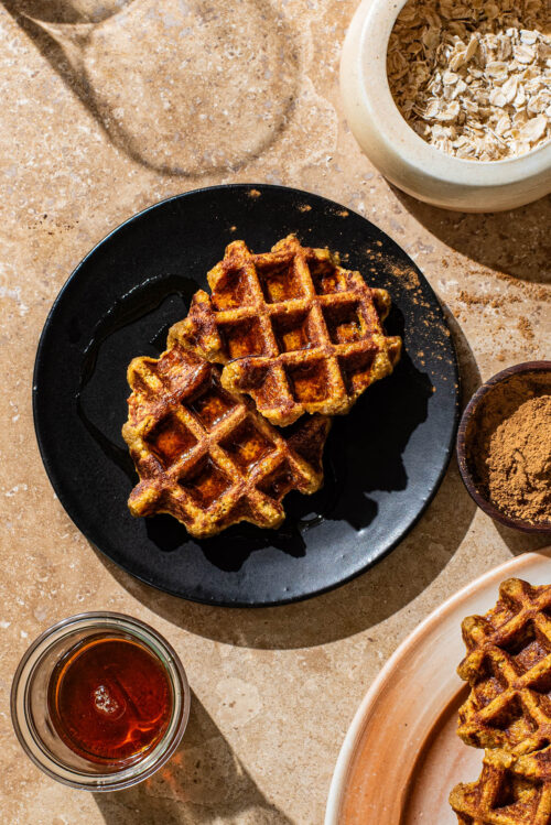 Two small round waffles on a black plate with more waffles and toppings around.