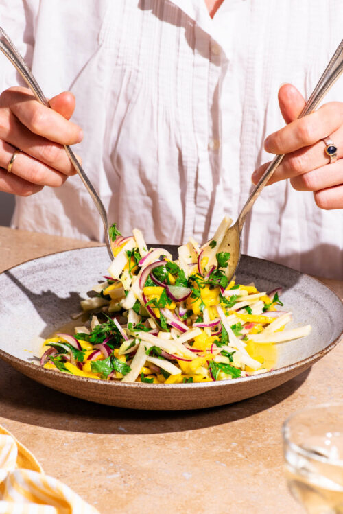 Woman's hands tossing a mango vegetable salad in a serving bowl.
