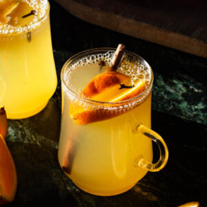 Two glasses of orange tea with pieces of orange and spices in them.