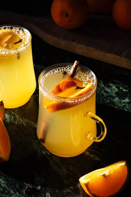 Two glasses of orange tea with pieces of orange and spices in them.