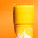 A lighter orange smoothie in a glass with yoghurt swirled at the base, against a darker orange background.