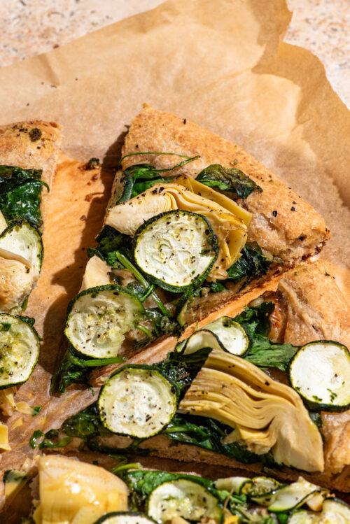 Slices of vegetable pizza with greens, artichoke hearts, and zucchini rounds.