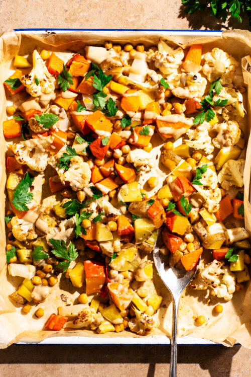 Vegetable tray bake with tahini sauce and parsley on the baking sheet with a serving spoon.