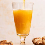 A bubbly golden drink being poured into a glass with ginger beside.