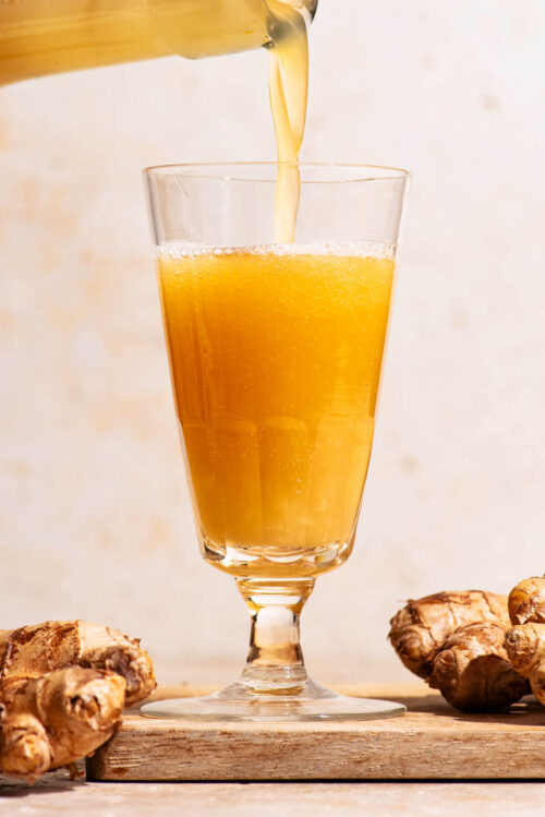 A bubbly golden drink being poured into a glass with ginger beside.
