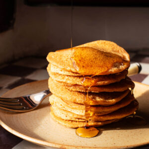 A stack of pancakes on a small plate, topped with maple syrup.