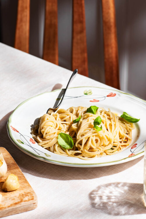 Spaghetti with a light sauce on a plate, topped with basil leaves, at a table setting.