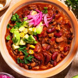 A bowl of bean chili topped with pickled onions, avocado, and herbs.
