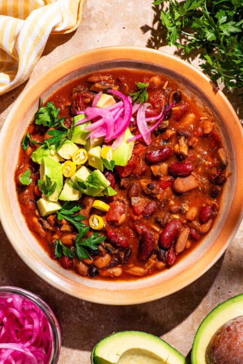A bowl of bean chili topped with pickled onions, avocado, and herbs.