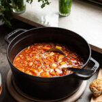 A large black pot filled with tomato vegetable soup in front of a windowsill.