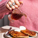 Pancakes on a plate topped with yogurt and a woman pouring maple syrup over them.