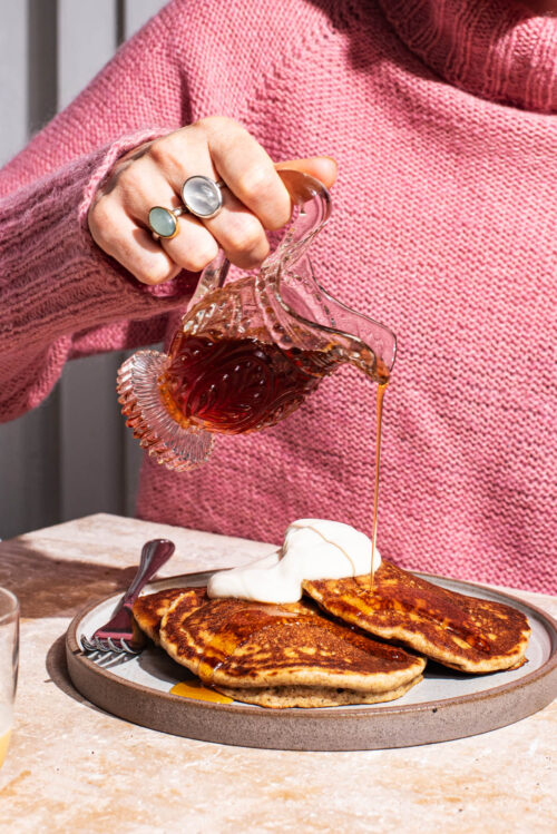 Pancakes on a plate topped with yogurt and a woman pouring maple syrup over them.