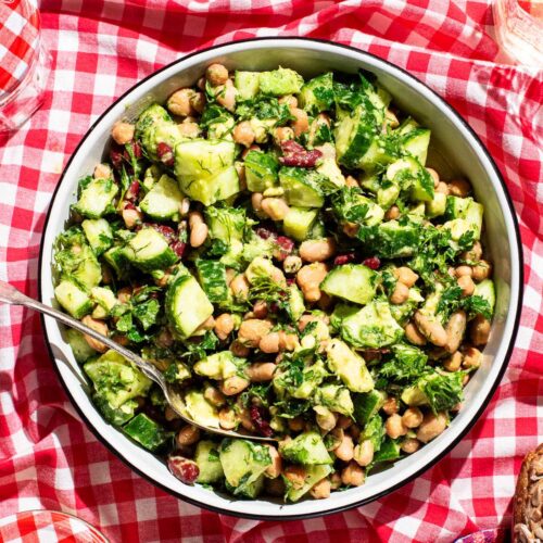 A large bowl of bean salad with cucumber and avocado on a red gingham picnic blanket.