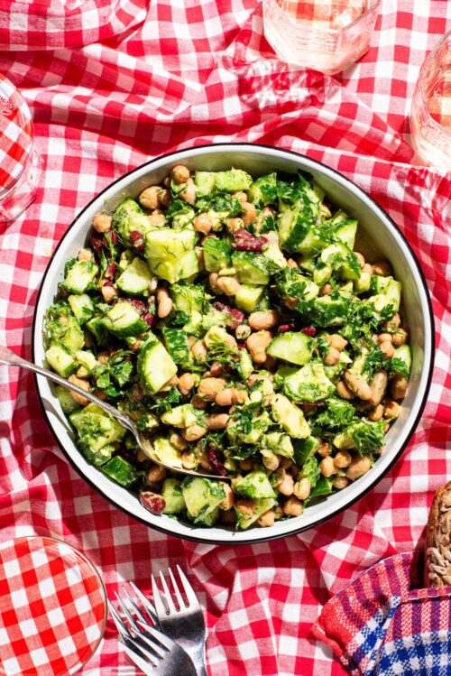 A large bowl of bean salad with cucumber and avocado on a red gingham picnic blanket.