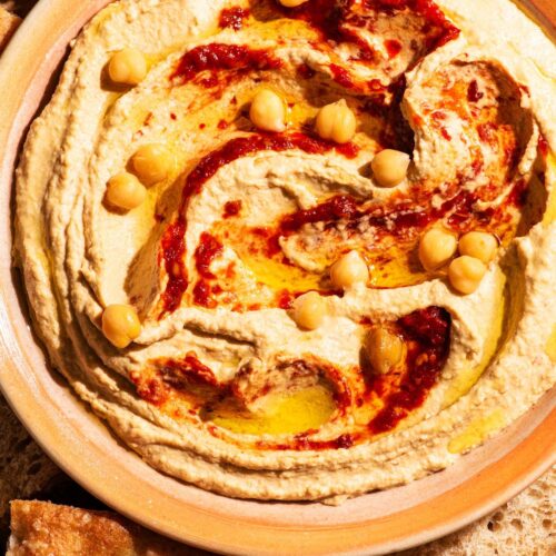 A bowl of hummus topped with hot pepper sauce and chickpeas, with bread around.