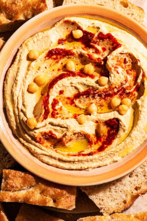 A bowl of hummus topped with hot pepper sauce and chickpeas, with bread around.