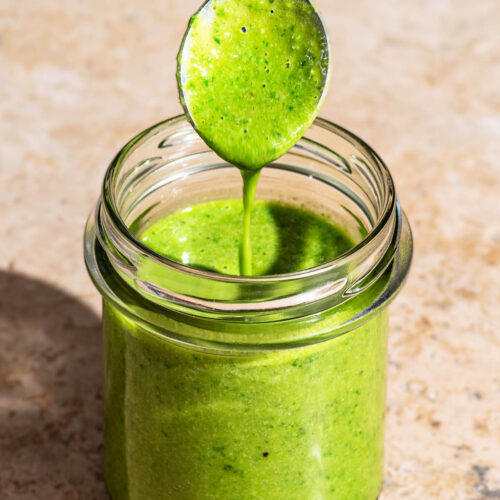 A glass jar filled with basil salad dressing, with a spoon dripping the dressing back in to the jar.