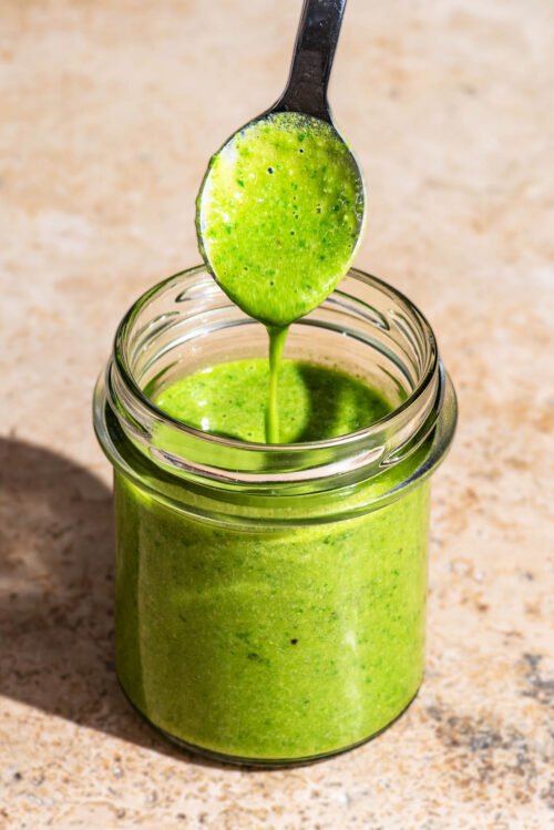 A glass jar filled with basil salad dressing, with a spoon dripping the dressing back in to the jar.
