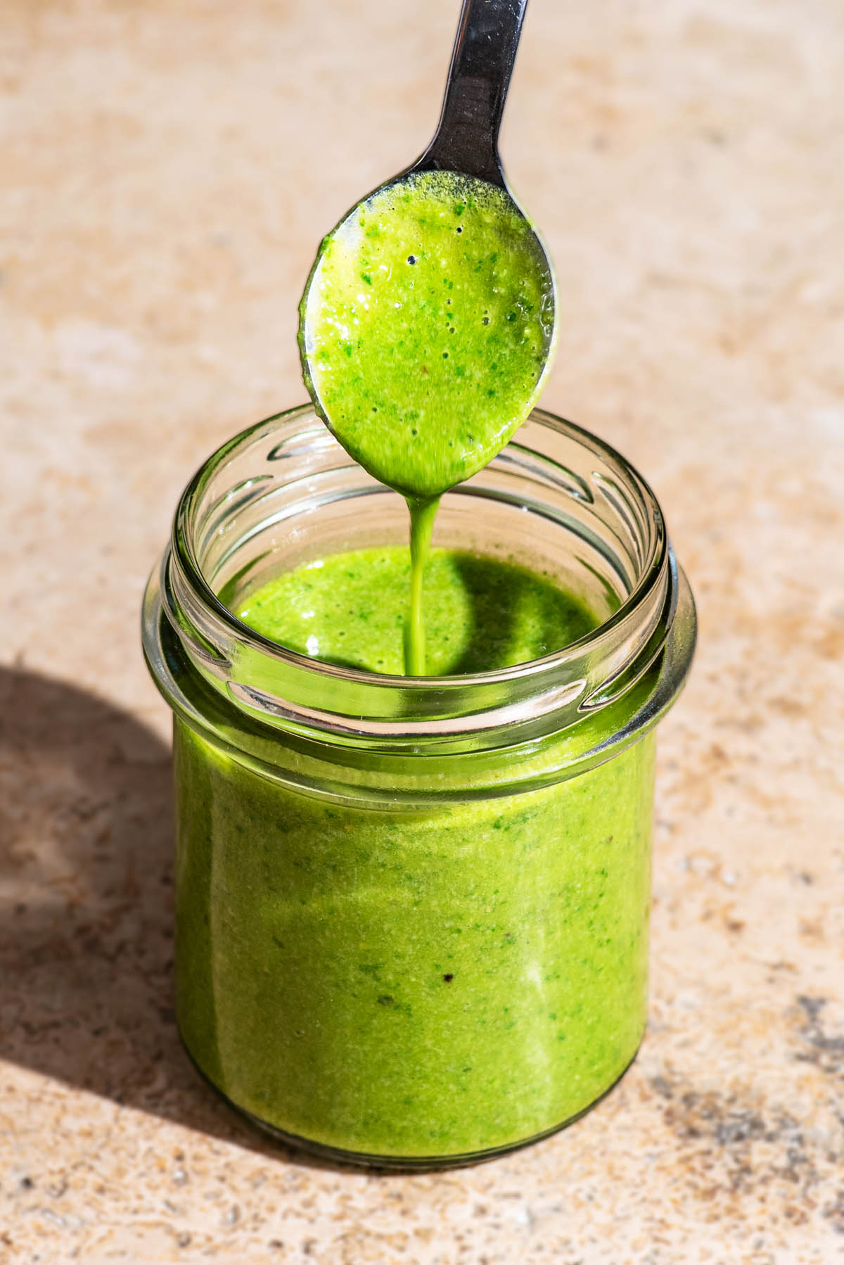 A glass jar filled with basil salad dressing, with a spoon dripping the dressing back in to the jar.