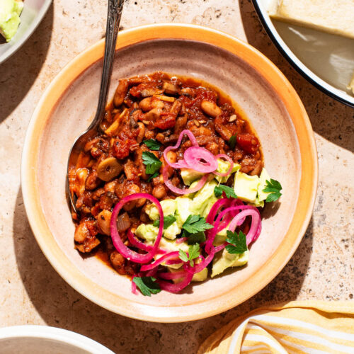 Bowls of chili topped with avocado and red onion, with buns beside.