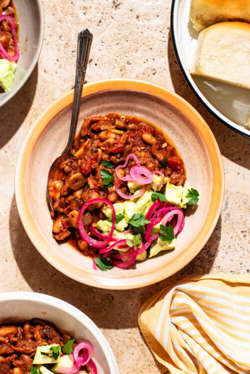 Bowls of chili topped with avocado and red onion, with buns beside.