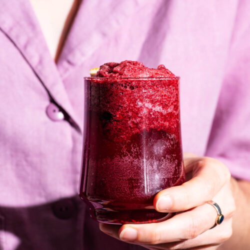 Woman holding a foamy purple drink and wearing a lighter purple blouse.