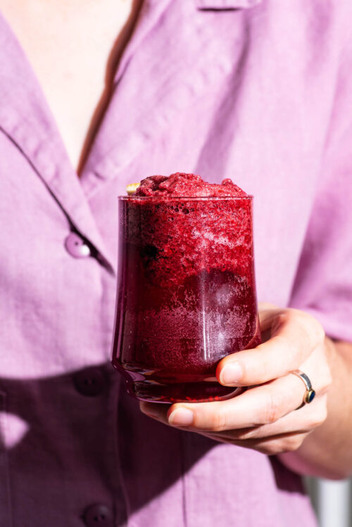 Woman holding a foamy purple drink and wearing a lighter purple blouse.