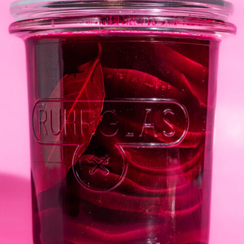 Glass jar filled with pickled beets on a pink background.