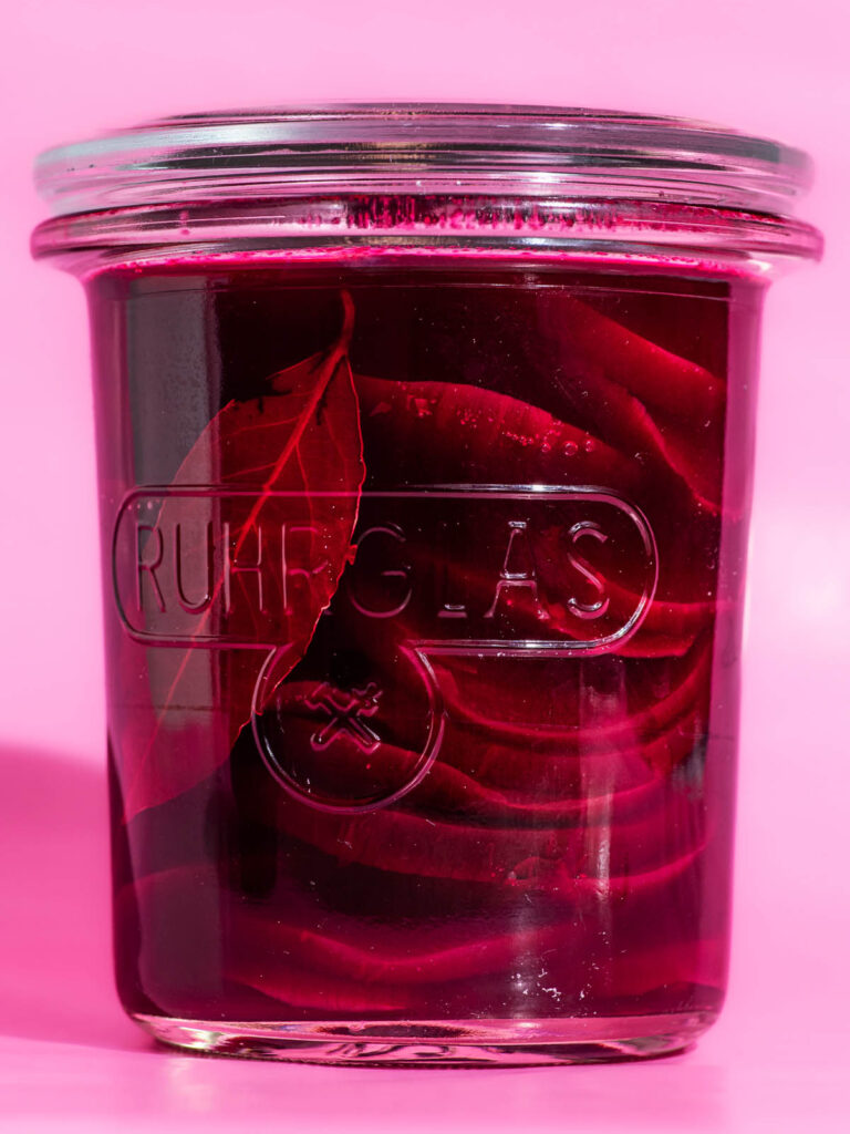 Glass jar filled with pickled beets on a pink background.