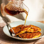 Pouring maple syrup over a plate of three orange pancakes on a green plate.