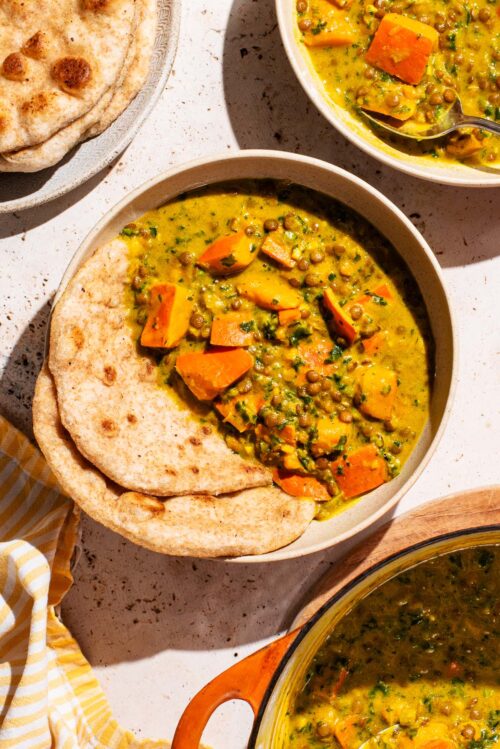 Two bowls of curry with flatbread and a pot in the corner.