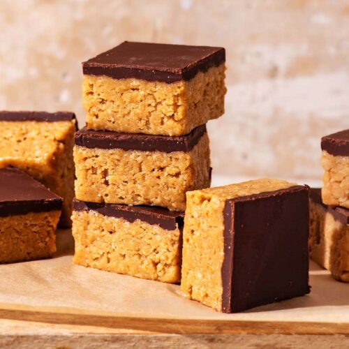 Several chocolate-topped peanut butter bars stacked and piled on a small cutting board.