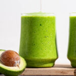 Green smoothie in two glasses with a halved avocado.