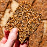 Seed-based cracker held in a hand above a tray with more crackers.