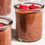 Small glass jars of chocolate chia seed pudding topped with raspberries.
