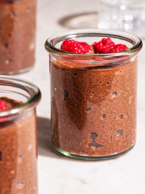 Small glass jars of chocolate chia seed pudding topped with raspberries.