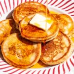 A red striped plate with six small pancakes on it, a piece of butter, and being drizzled with syrup.