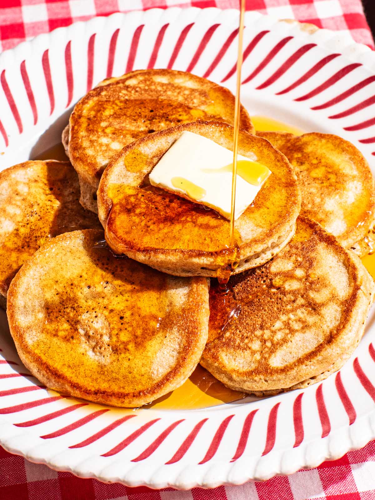 A red striped plate with six small pancakes on it, a piece of butter, and being drizzled with syrup.