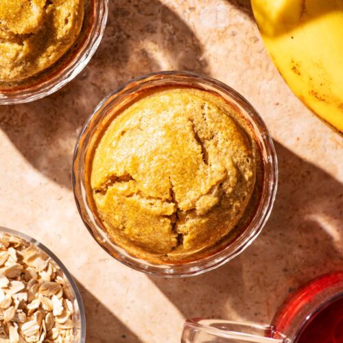 Two servings of baked oats in glass dishes with banana, tea, and rolled oats around.