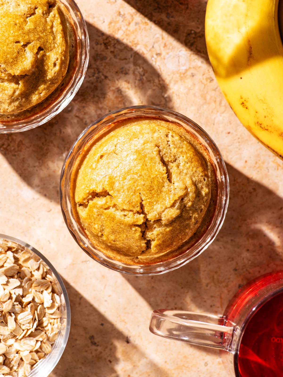 Two servings of baked oats in glass dishes with banana, tea, and rolled oats around.