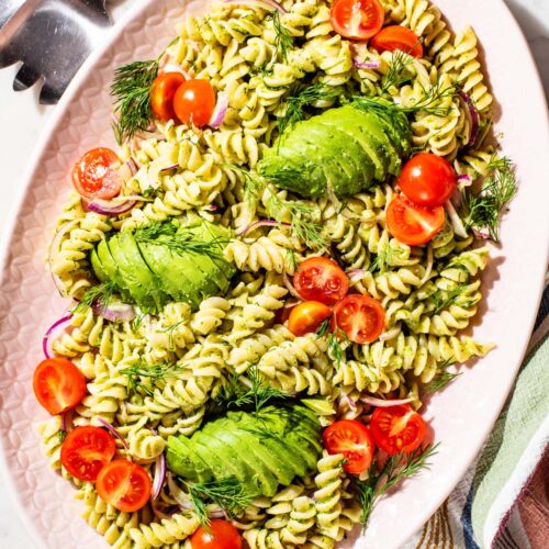 Pasta salad on a large platter with sliced avocado, cherry tomatoes, and red onion.