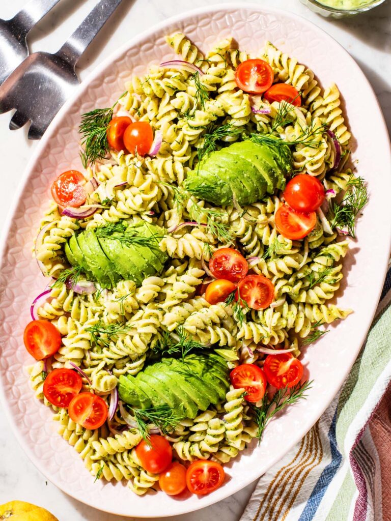 Pasta salad on a large platter with sliced avocado, cherry tomatoes, and red onion.