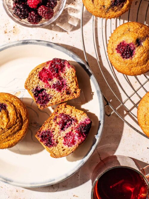 Two blackberry muffins on a plate, one halved, with more on a wire rack.