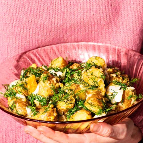 Woman in a pink sweater holding a pink glass serving bowl filled with potato salad.