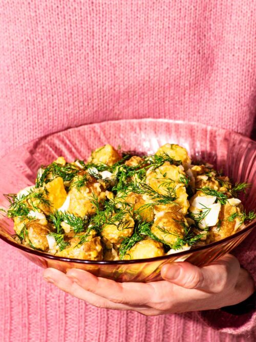 Woman in a pink sweater holding a pink glass serving bowl filled with potato salad.