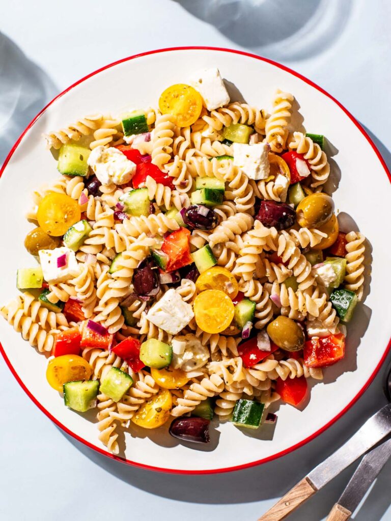 Pasta salad with olives, tomatoes, cucumber, and feta on a large serving plate.