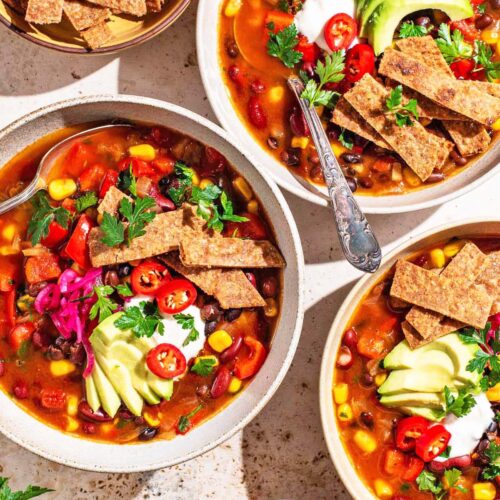 Three bowls of tomato-based soup topped with avocado, tortilla chips, and herbs.