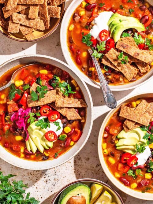 Three bowls of tomato-based soup topped with avocado, tortilla chips, and herbs.