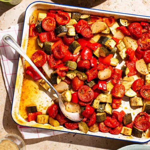 Roasted summer vegetables on a baking sheet with bowls and glasses around.